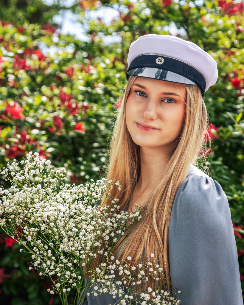 Nuori nainen hymyilee ylioppilaslakki päässä, kädessään valkoisia kukkia, taustalla kesäinen kukkiva puisto.