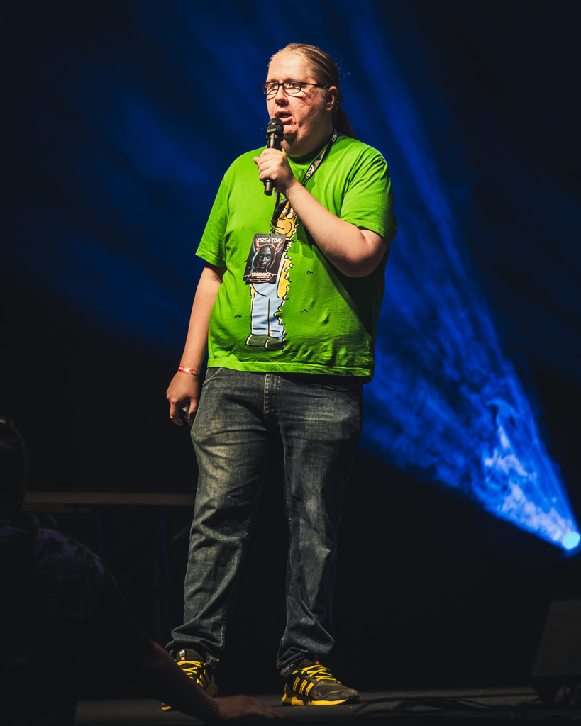 Assembly Summer tapahtuman juontaja lavalla vihreässä t-paidassa ja mikrofonin kanssa. Paulin pelivideot.