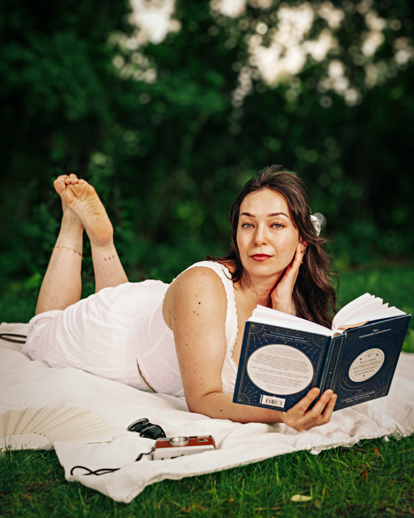 Henkilö istuu piknikviltillä ja lukee kirjaa kesäisessä puistossa, vierellä laukku ja sandaalit.