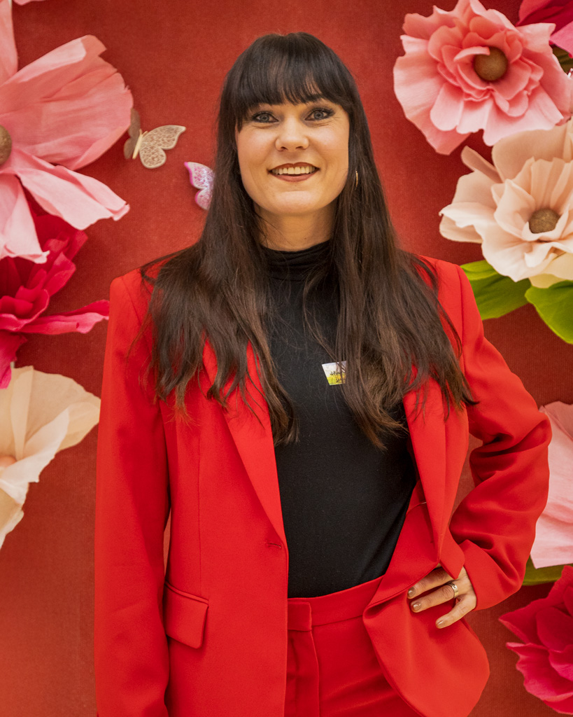 Mira Luoti poseeraa punaisessa puvussa kukkakoristeisen taustan edessä yrittäjyystapahtumassa.