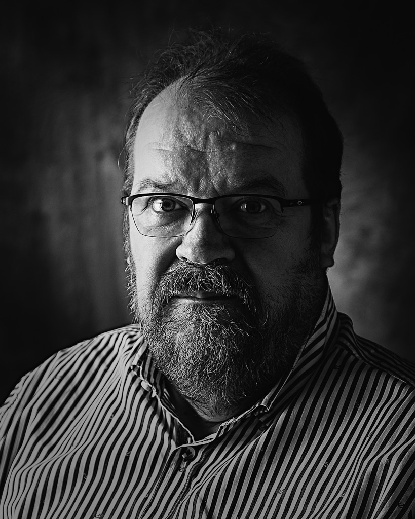 Mustavalkoinen muotokuva, mies katsoo kameraan tummassa taustassa, studiossa otettu henkilökuva.