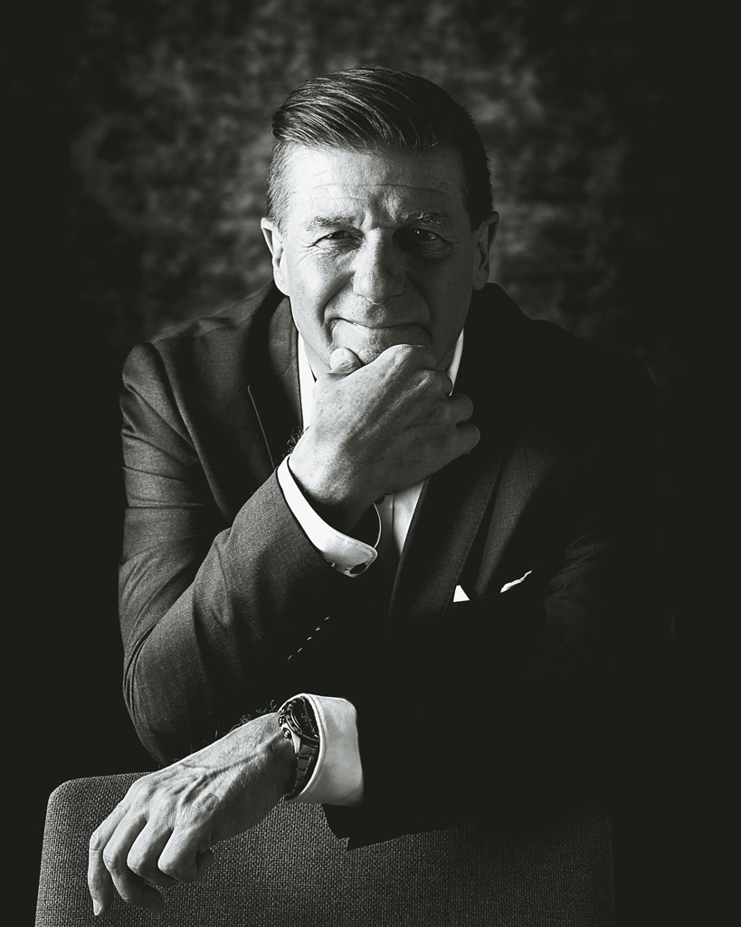 Mustavalkoinen henkilökuva, mies istuu nojaten käteen ja katsoo kameraan itsevarmasti, studioympäristö.