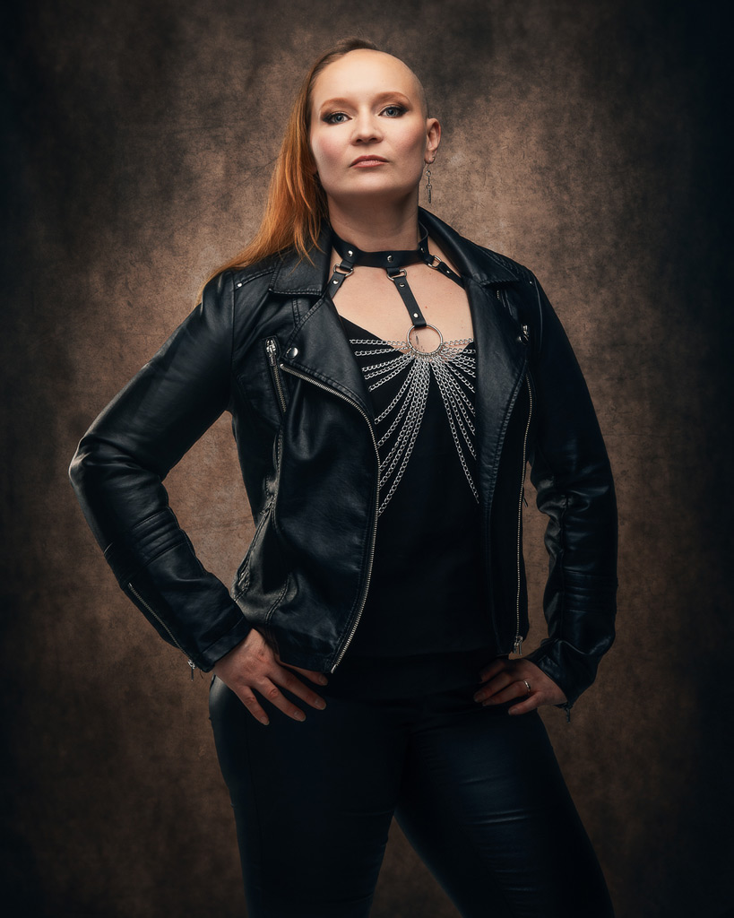 Henkilö seisoo kädet lanteilla ja katsoo kameraan itsevarmasti, musta nahkatakki ja tummat vaatteet, taustalla ruskea studiotausta.