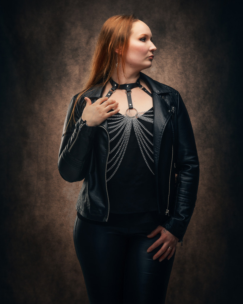 Henkilö seisoo sivuttain katse suunnattuna vasemmalle, pukeutuneena mustaan nahkatakkiin ja ketjukoristeiseen yläosaan, taustalla ruskea studiotausta.