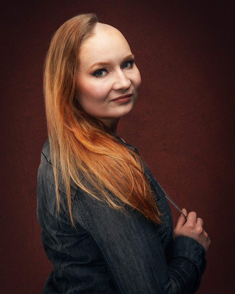 Henkilö katsoo olkansa yli hymyillen, denimtakki ja pitkät hiukset, taustalla punaruskea studiotausta.