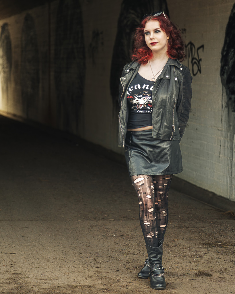 Henkilö seisoo nahkatakissa ja mustassa asussa, taustalla alikulkukäytävä ja seinämaalauksia.